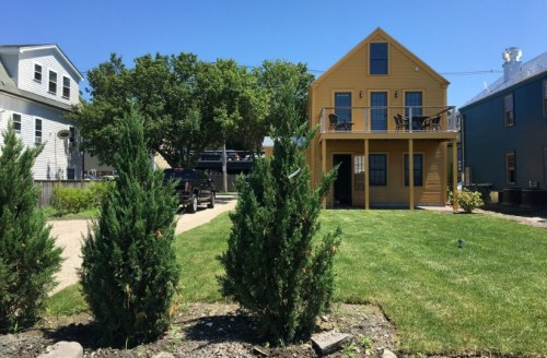 A yellow two-story house with a balcony, surrounded by green grass and small trees, under a clear blue sky.