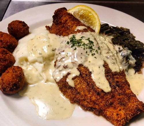 A plate of fried fish topped with creamy sauce, served with mashed potatoes, collard greens, and fried cornmeal balls.