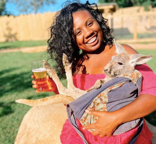 A smiling woman in a pink dress holds a kangaroo in a pouch while enjoying a drink outdoors.