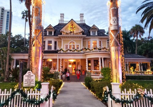 A beautifully decorated historic house for the holidays, adorned with lights and festive decorations, surrounded by palm trees.