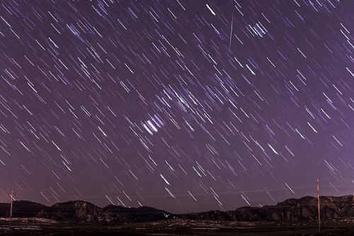 A long-exposure shot of a starry night sky, showing streaks of light against a purple backdrop and silhouetted mountains.