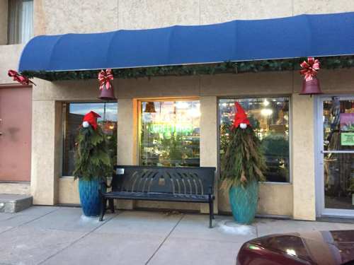 A festive storefront decorated with greenery, red bows, and Santa hats on planters, featuring a bench outside.