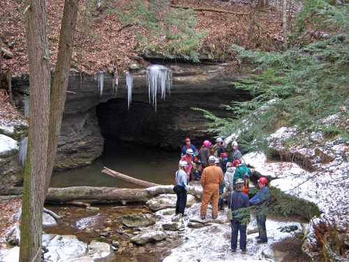 A group of people in helmets gathers near a cave entrance surrounded by snow and icicles in a wooded area.