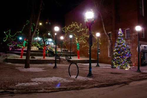 A festive park at night, adorned with colorful Christmas lights and a decorated tree, surrounded by snow.