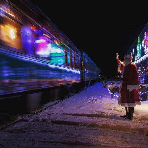 A Santa figure stands on a snowy platform as a brightly lit train passes by at night.