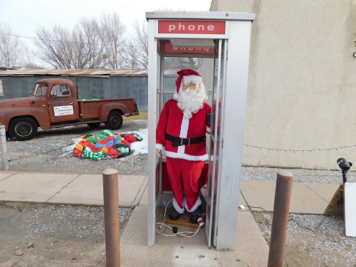 A Santa Claus figure stands in a phone booth, with a vintage truck and holiday decorations in the background.
