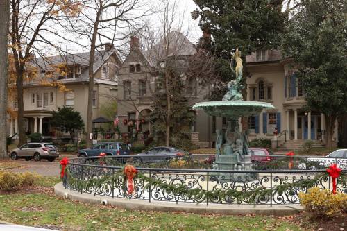 A decorative fountain surrounded by festive greenery and historic homes, with cars parked nearby on a cloudy day.