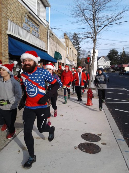 A group of runners in Santa hats and festive attire jogs down a sidewalk during a holiday event.