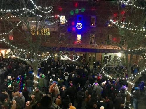 A large crowd gathers outdoors, surrounded by festive lights and decorations, celebrating in a lively atmosphere.