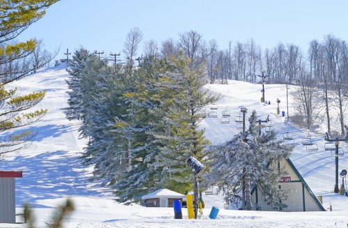 A snowy ski slope with chairlifts, surrounded by trees and a clear blue sky.