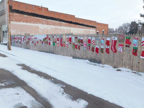 A snowy street scene featuring a wooden fence adorned with colorful Christmas stockings.