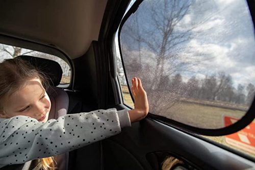 A young girl smiles while reaching out to a car window, with trees and a cloudy sky visible outside.