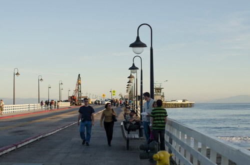 A scenic pier with people walking, sitting on benches, and enjoying the ocean view under street lamps.