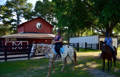 Two riders on horses in front of a red barn, surrounded by trees and a fenced area.