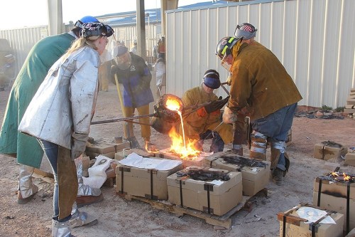 Workers in protective gear pour molten metal into molds at a foundry, surrounded by smoke and equipment.