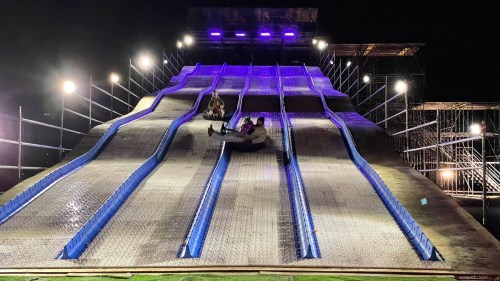 Two people sliding down a large, illuminated water slide at night, with multiple lanes and bright lights in the background.