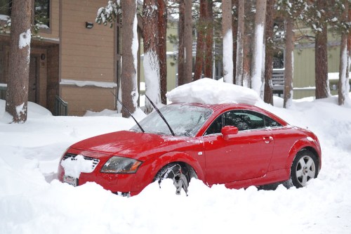 A red car partially buried in snow, with snow piled on the roof and wipers raised, surrounded by snowy trees.