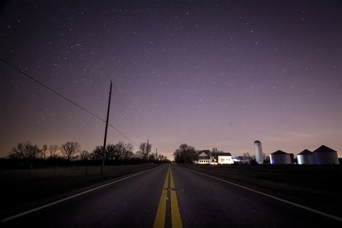 A quiet rural road at night, lined with trees, leading to a farm with silos under a starry sky.