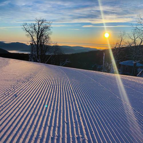 A serene winter landscape with freshly groomed snow, trees, and a vibrant sunrise over distant mountains.