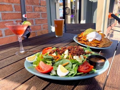 A vibrant salad with eggs and vegetables, a plate of nachos, a cocktail, and a beer on a wooden table outdoors.