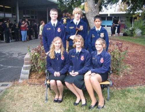 A group of six students in blue jackets poses for a photo outside a school building, with trees and people in the background.