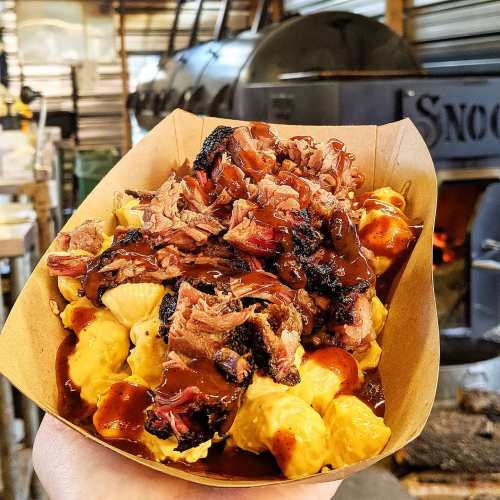 A hand holding a tray of macaroni and cheese topped with pulled pork and barbecue sauce, set against a smoky background.