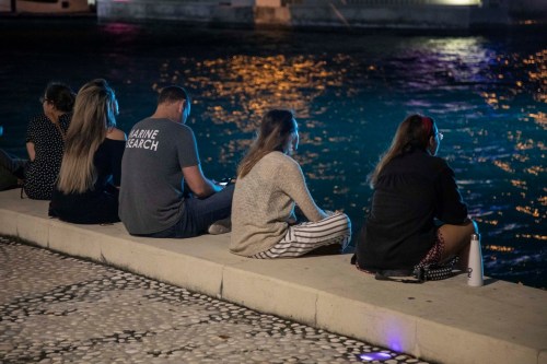 A group of people sitting on a waterfront at night, gazing at the water illuminated by colorful lights.
