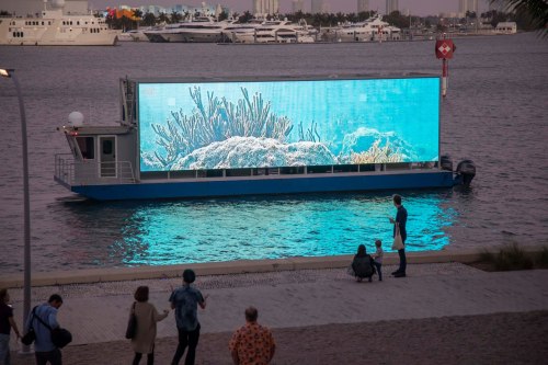 A boat with a large digital screen displays vibrant underwater coral scenes, with people observing from the shore.