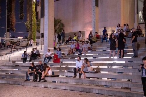 A diverse crowd sits on outdoor steps, some chatting and others looking at their phones, under evening lights.