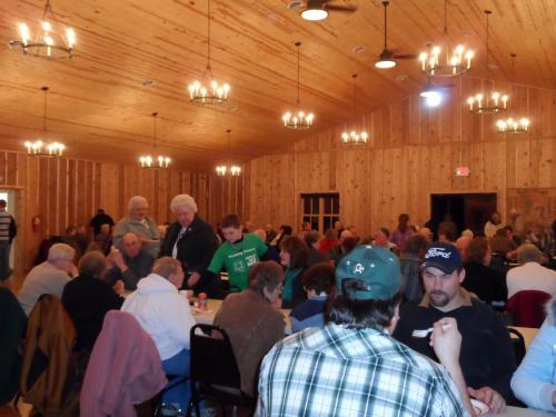 A crowded indoor hall with wooden walls, people seated at tables, chatting and enjoying a gathering.