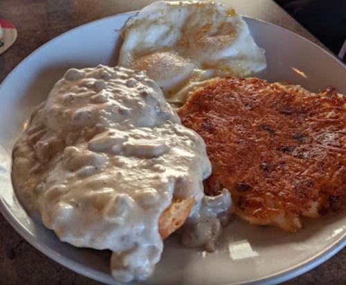 A plate featuring biscuits smothered in gravy, two fried eggs, and a crispy potato pancake.