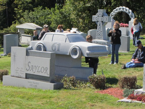 A unique gravestone shaped like a car, surrounded by visitors in a cemetery with various headstones.