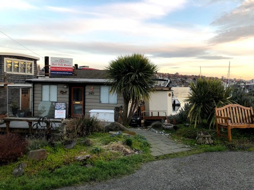A cozy waterfront house with a sign, surrounded by greenery and a bench, under a colorful sky.