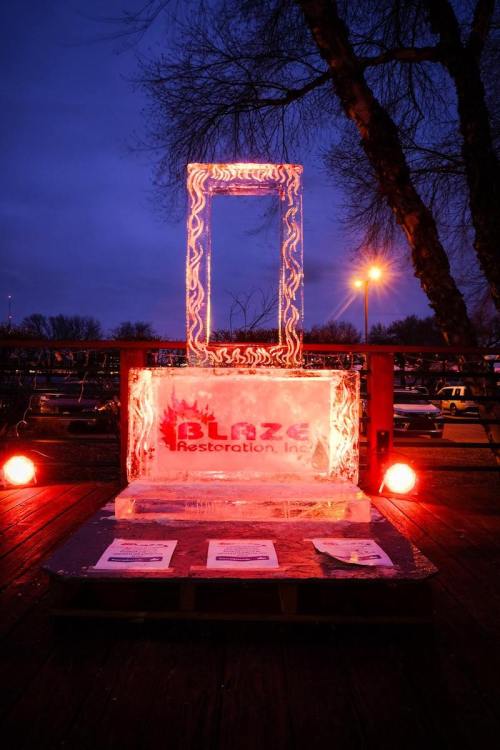 An illuminated ice sculpture with a rectangular frame, featuring the logo of Blaze Restoration, set against a twilight sky.