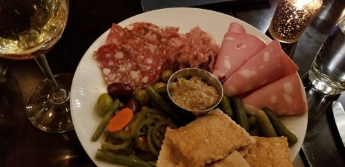 A plate of assorted meats, pickles, olives, and crackers, accompanied by a glass of white wine.