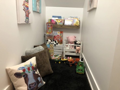 Cozy play nook with toys, plush animals, and colorful books, featuring a cow-patterned pillow and soft carpet.