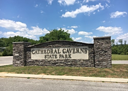 Sign for Cathedral Caverns State Park, featuring stone pillars and a blue sky with scattered clouds in the background.