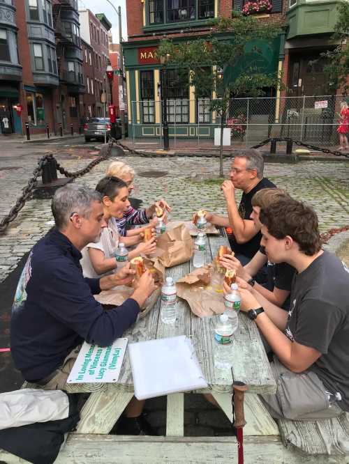 A group of six people sitting at a picnic table, enjoying food and drinks in an outdoor urban setting.