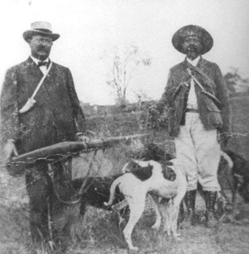 Two men in vintage clothing stand with hunting rifles and dogs in a rural setting.
