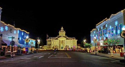 A festive town square at night, adorned with colorful holiday lights and decorations on buildings and trees.
