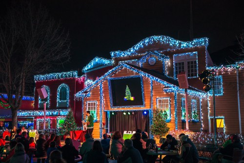A festive outdoor scene with a decorated building, colorful lights, and an audience enjoying a performance.
