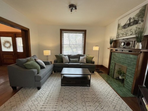 Cozy living room with gray sofas, a coffee table, and a green-tiled fireplace, featuring natural light and decorative plants.