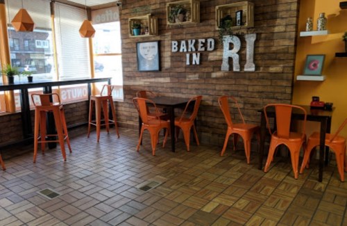 Cozy café interior with orange chairs, wooden walls, and a "Baked in RI" sign. Bright and inviting atmosphere.