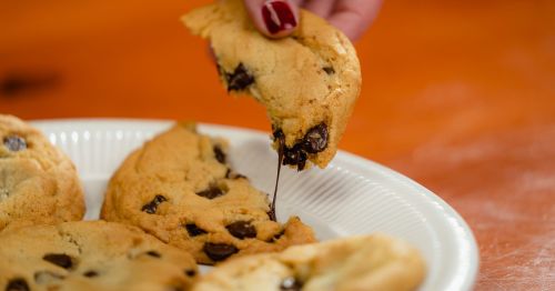 A hand reaches for a chocolate chip cookie, with gooey chocolate stretching from the cookie on a white plate.