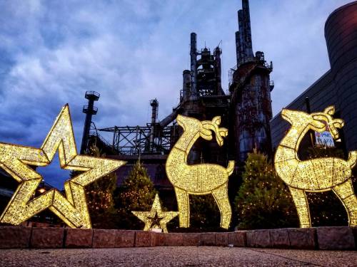 Illuminated holiday decorations, including reindeer and stars, in front of an industrial backdrop with smokestacks.