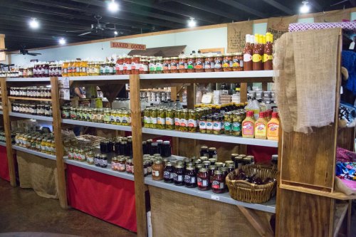 A display of various jars and bottles of sauces, jams, and pickles on wooden shelves in a market setting.