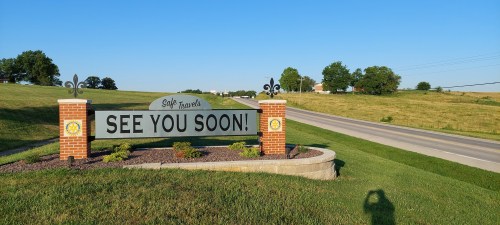 A sign reading "See You Soon!" stands by a road, surrounded by green grass and trees under a clear blue sky.