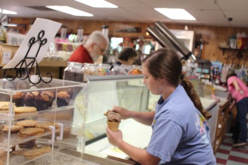 A young woman scoops ice cream into a cone at a busy ice cream shop, with customers in the background.