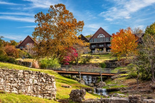 A scenic view of a lodge surrounded by colorful autumn trees and a small waterfall, with a bridge in the foreground.