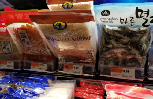 Packages of dried seafood, including small shrimp and anchovies, displayed on a grocery store shelf with price tags.